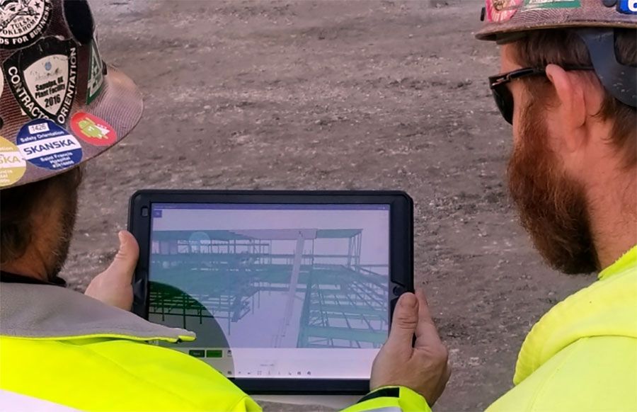Two construction workers in hard hats and high-visibility vests look at a tablet displaying a 3D building model.