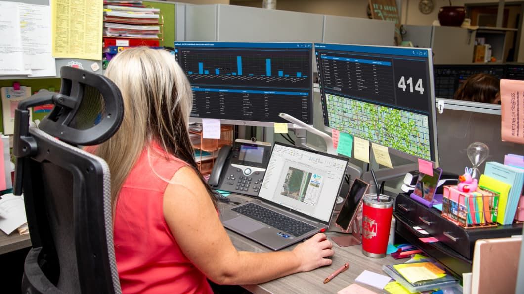 Person in red top working at desk with multiple monitors displaying data charts, maps, and analytics in office setting.