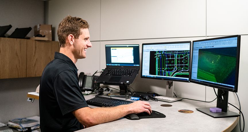 Man smiling while working on CAD software on a multi-monitor setup.