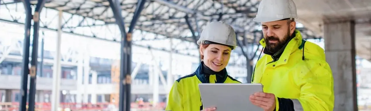 Two construction workers in yellow jackets and hard hats review plans on a tablet at a building site.
