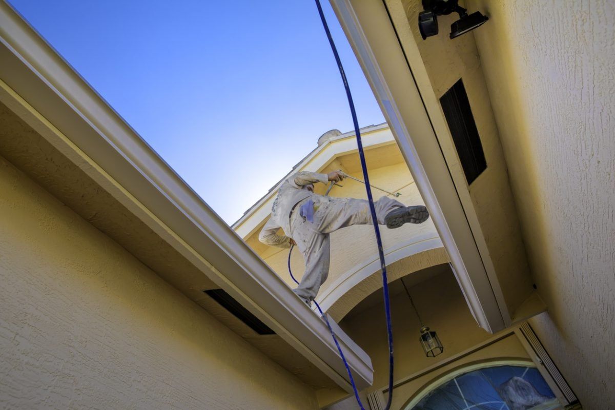 Low-angle view of a painter in paint-splattered clothes spray painting a building's exterior against a blue sky.