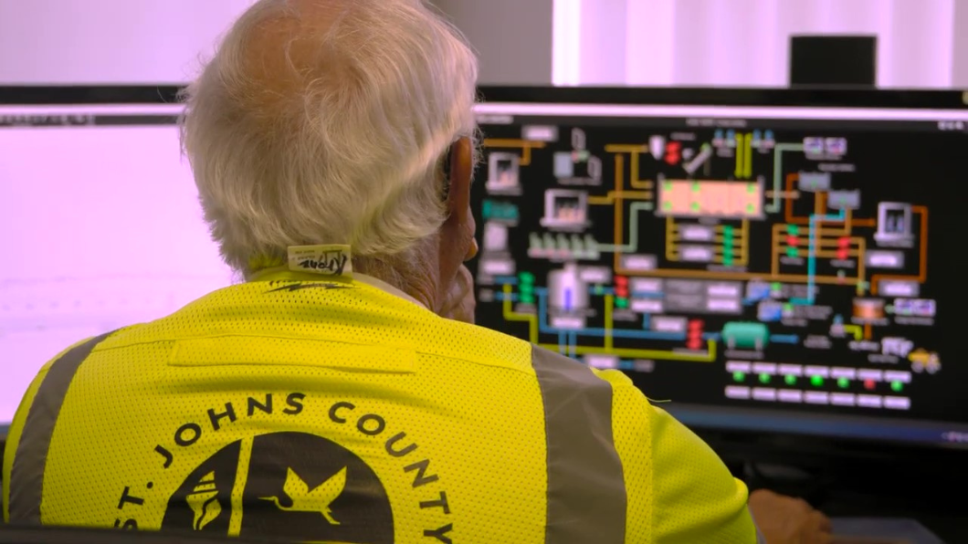 A man wearing a St. Johns County safety vest monitors a complex, colorful schematic diagram of a utility system on a large computer screen.