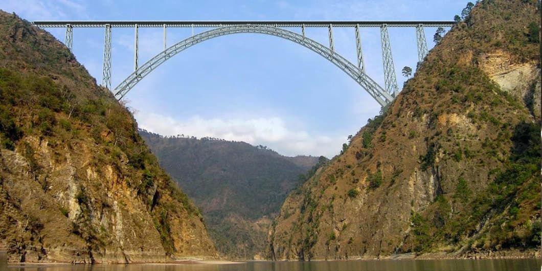 A steel truss arch bridge spans a wide river valley between two steep, rocky mountainsides.