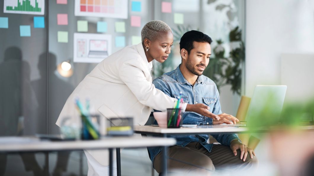 Colleagues collaborating at a desk with a laptop and sticky notes on the wall.