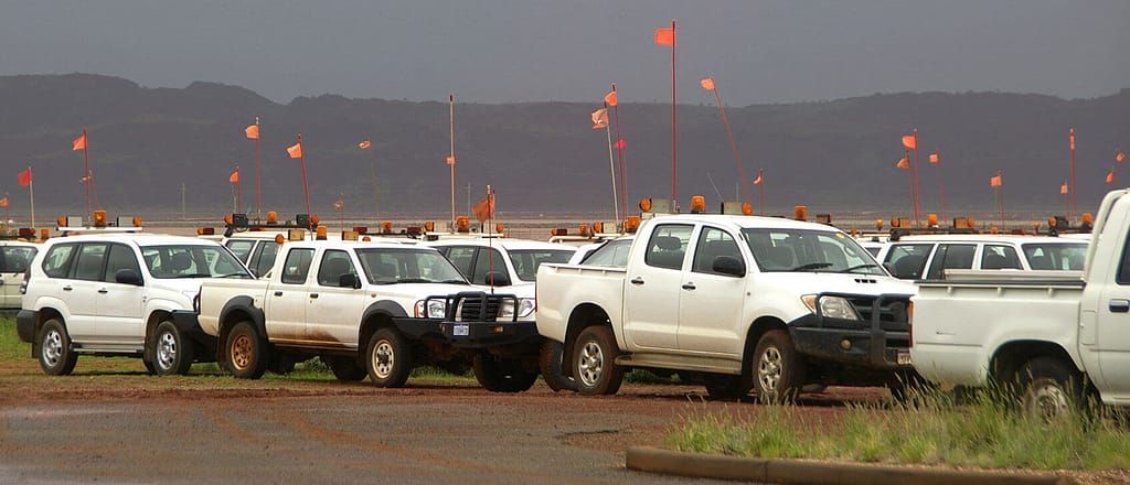 A fleet of white trucks and SUVs are parked in a field with orange flags on poles in the background.