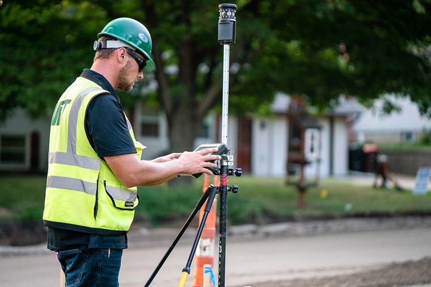 Construction worker in a hard hat and safety vest operates surveying equipment on a tripod outdoors.