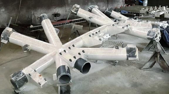A white, star-shaped structure made of large, cylindrical metal tubes is partially wrapped in plastic sheeting, in a workshop environment.