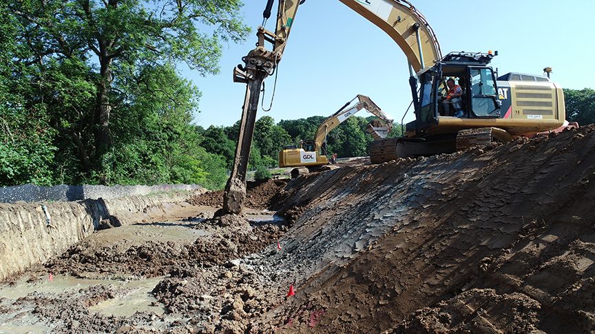 Excavators dig a trench on a sunny day with trees in the background.