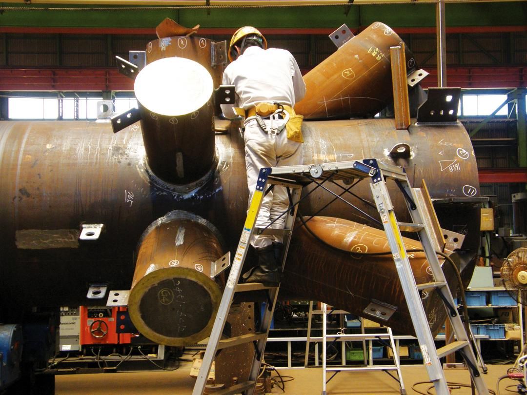A worker in a hard hat and white overalls stands on a ladder assembling a large, complex metal structure in a factory.