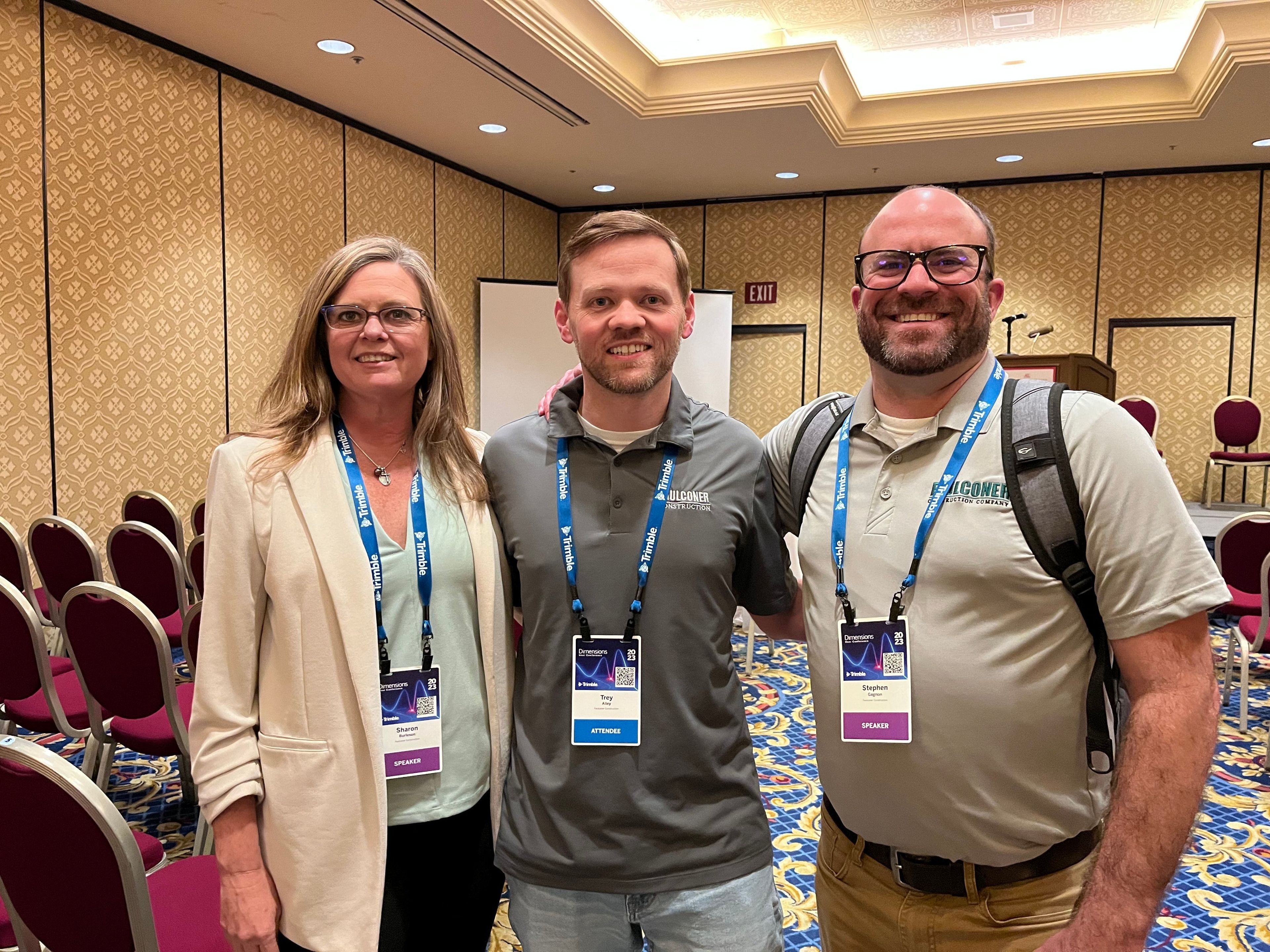 Three individuals, pose for a photo in front of a conference room backdrop, all wearing lanyards with name tags.