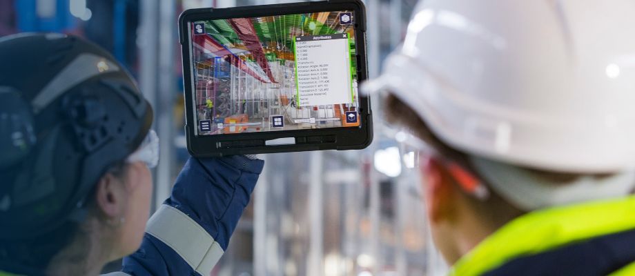 Two construction workers in hard hats and safety vests look at a tablet displaying a 3D model of a construction site.