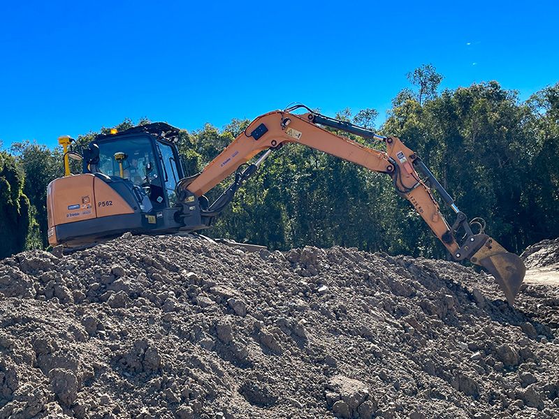 Orange excavator on a pile of dirt against a blue sky and trees.