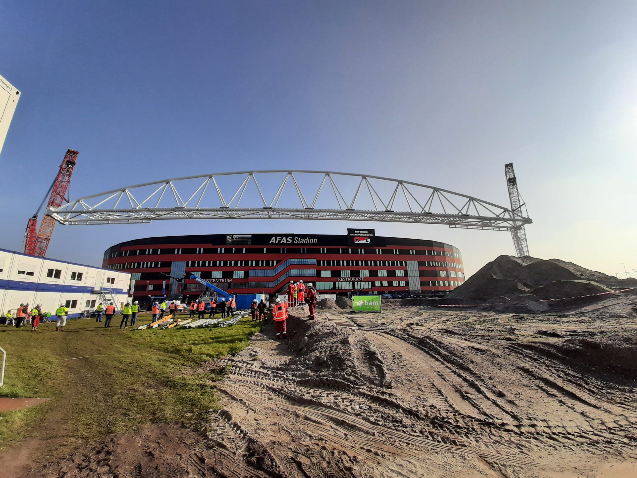 Construction workers in hard hats gather near a large stadium under construction, with a massive steel truss being lifted into place by cranes.