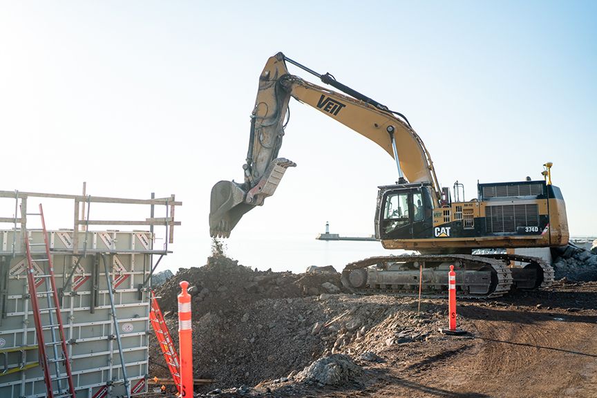 Yellow excavator working on a construction site near the water, with a lighthouse in the distance.