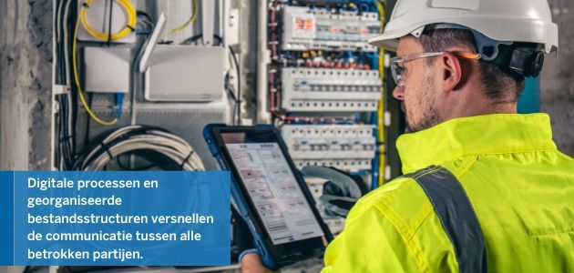 Technician in a hard hat and hi-vis vest examining a tablet in front of an electrical panel.
