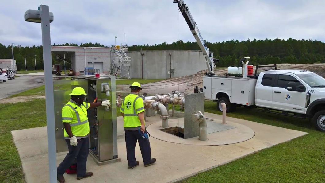 Two utility workers in yellow safety vests and hard hats inspect a control panel at an industrial site.