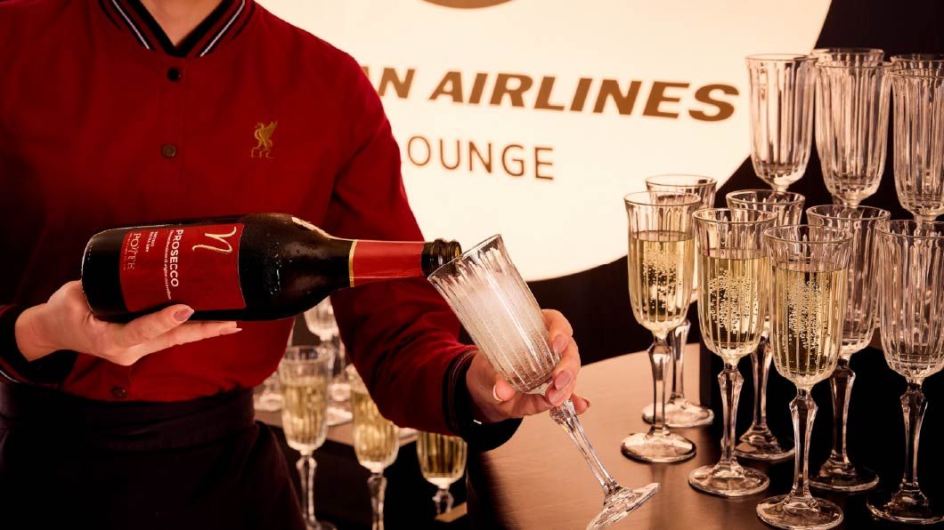 A staff member in red pours prosecco into a flute at an airline lounge, with multiple crystal champagne glasses on the counter.