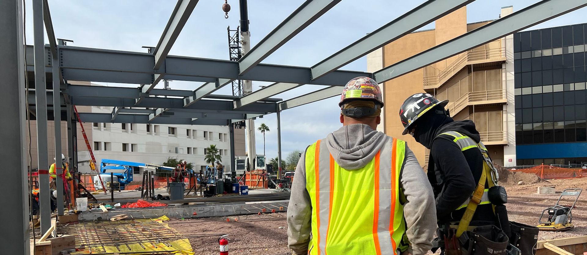 Two construction workers in hard hats and safety vests observe the progress of a large steel building framework on a busy job site.