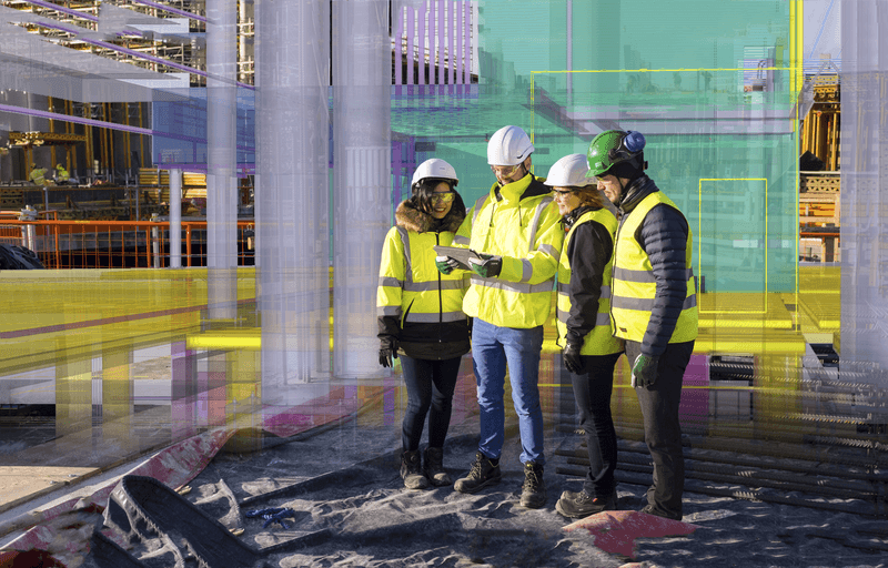 Four construction workers in hard hats and safety vests look at a tablet displaying a digital model of a building overlayed on a construction site.