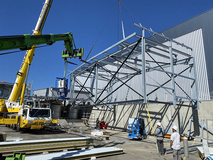 Workers use a crane and a lift to install a steel structure next to a modern building.