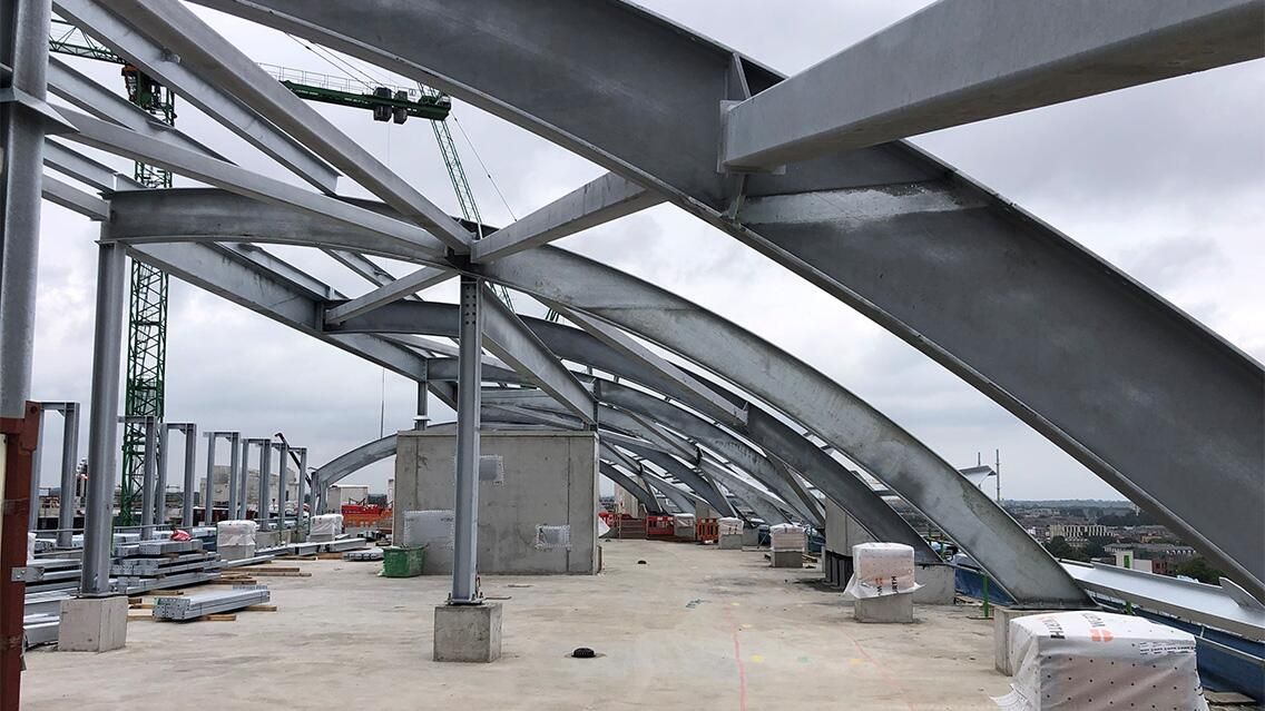 Close-up of a building's internal steel structure during construction, with arched beams and support columns visible against a cloudy sky.
