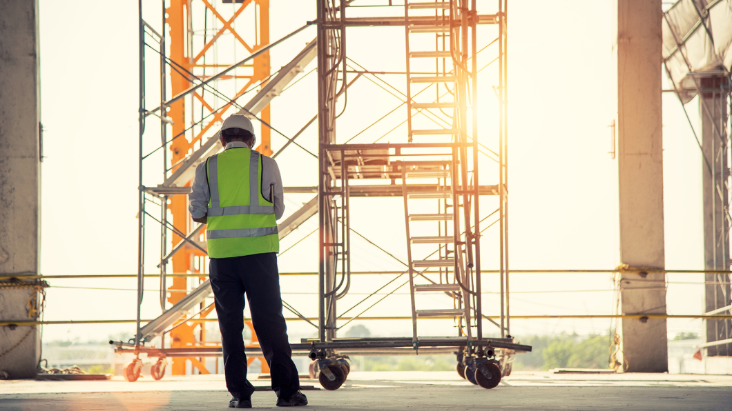 Construction worker in safety gear stands near scaffolding at a building site during sunset.